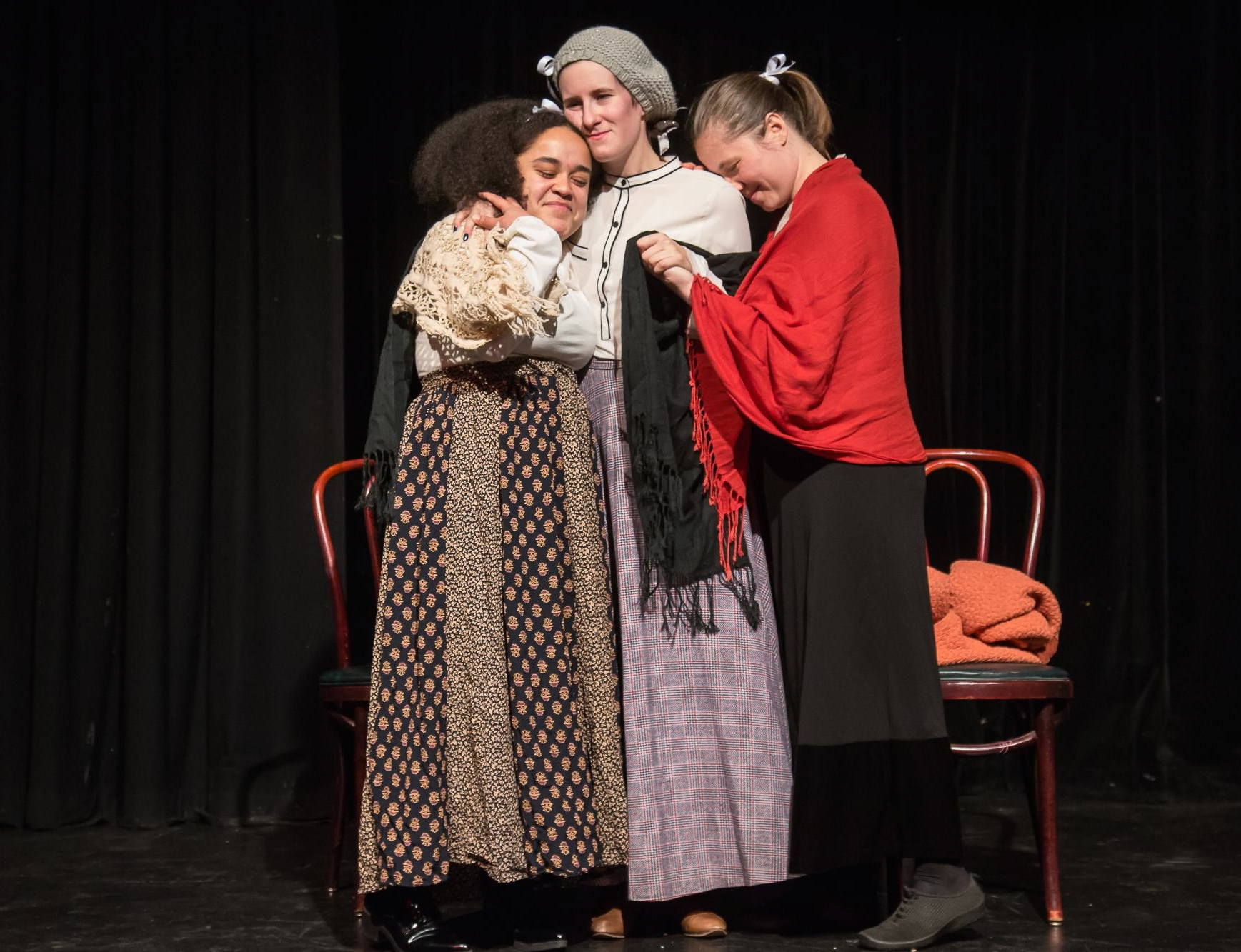 Three characters, who appear to be adult women dressed in Little Women-inspired dress, group hug